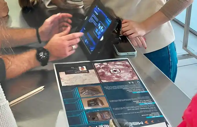 Mão a tocar no iPad com realidade aumentada, ao lado da ficha da missão, num escape game em sala