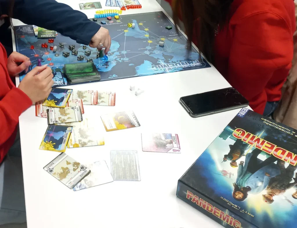 Jogadores a mover peças e cartas num jogo de tabuleiro cooperativo, sobre uma mesa branca