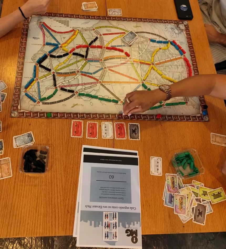 Mesa com um jogo de tabuleiro de comboios, cartas e a ficha de exercício da Outside Mind Games sobre o elevator pitch