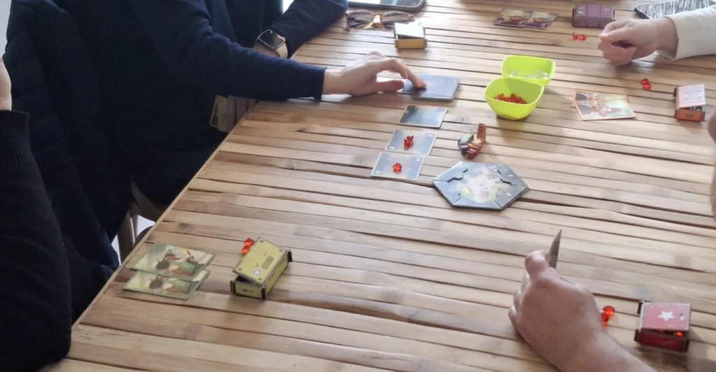 Mãos de jogadores à volta de uma mesa com um jogo de tabuleiro moderno, cartas e peças